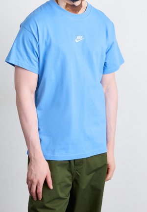 Hellblaues Baumwoll-T-Shirt mit Rundhalsausschnitt, kurzen Ärmeln und weißem Nike-Swoosh-Logo auf der Brust. Kombiniert mit grünen Hosen.