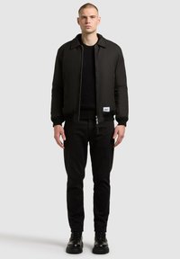 Schwarze Bomberjacke mit Kragen, Reißverschluss vorne und umgeschlagenen Ärmeln, getragen über einem schwarzen Sweater und engen schwarzen Hosen, kombiniert mit schwarzen Stiefeln.