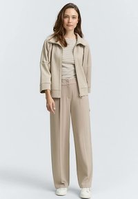 Veste de sport beige clair à col montant, associée à une chemise rayée et un pantalon beige ample. Des baskets blanches complètent la tenue.