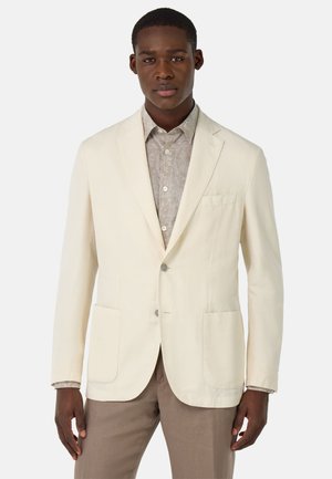 Blazer de algodón crema con dos bolsillos frontales, cierre de un solo botón, solapas con muesca y una textura suave, usado sobre una camisa estampada.