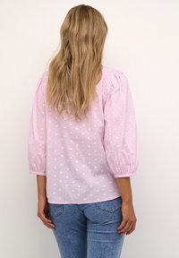 Blouse rose clair avec des manches bouffantes trois-quarts, texturée avec des points en relief. Associée à un jean bleu, montrant un design de tenue décontractée.