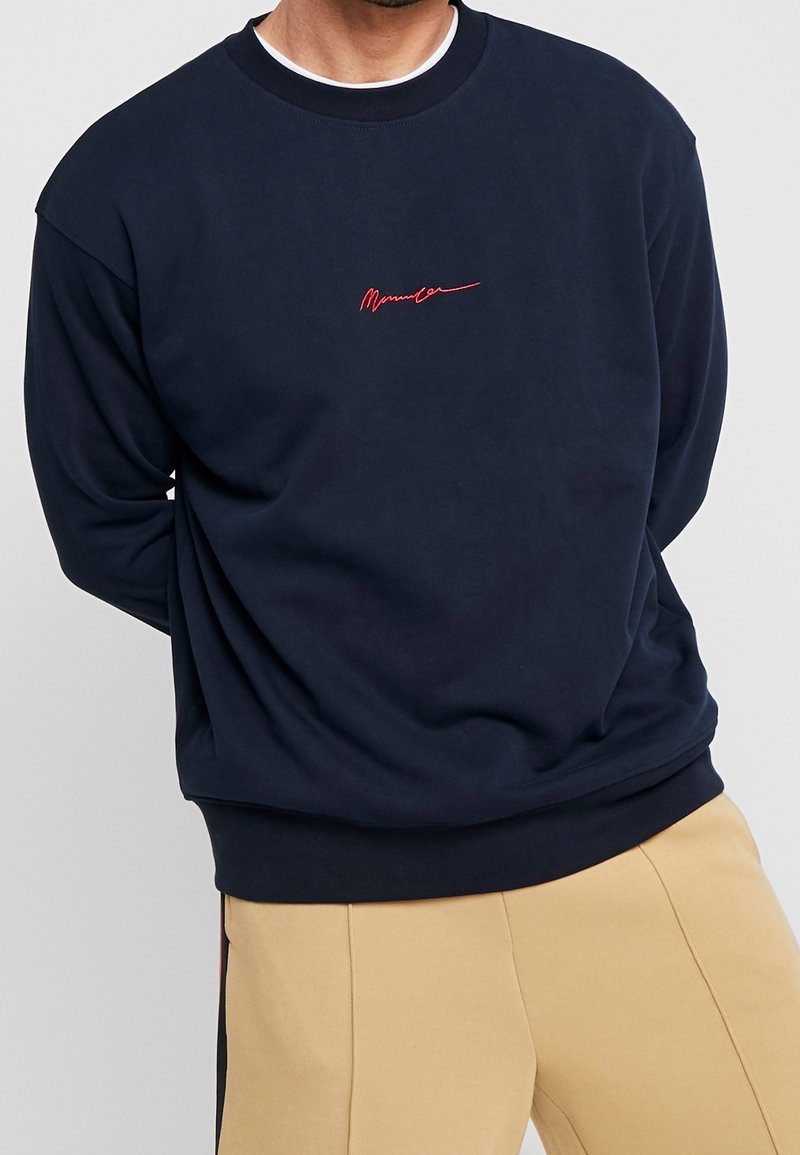 Homme portant un sweat-shirt bleu marine avec un logo rouge en écriture cursive au centre de la poitrine et un pantalon beige, mains derrière le dos, debout devant un fond uni.