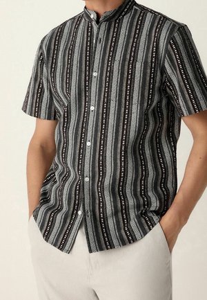Homme portant une chemise à manches courtes rayée verticalement noire et blanche avec un col mandarin et un pantalon clair.