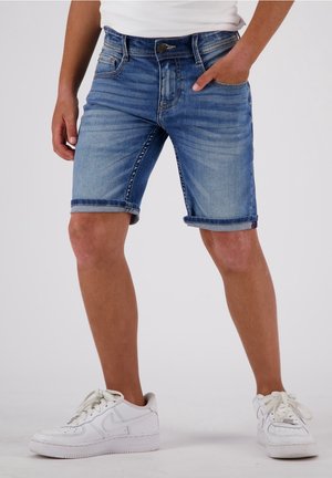 Personne portant un short en jean retroussé et des baskets blanches, debout avec une main dans la poche sur un fond uni.