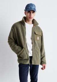 Giacca in pile verde oliva peloso con colletto alto, chiusura con zip e tasca sul petto; presenta dettagli in tessuto a contrasto e una vestibilità morbida.