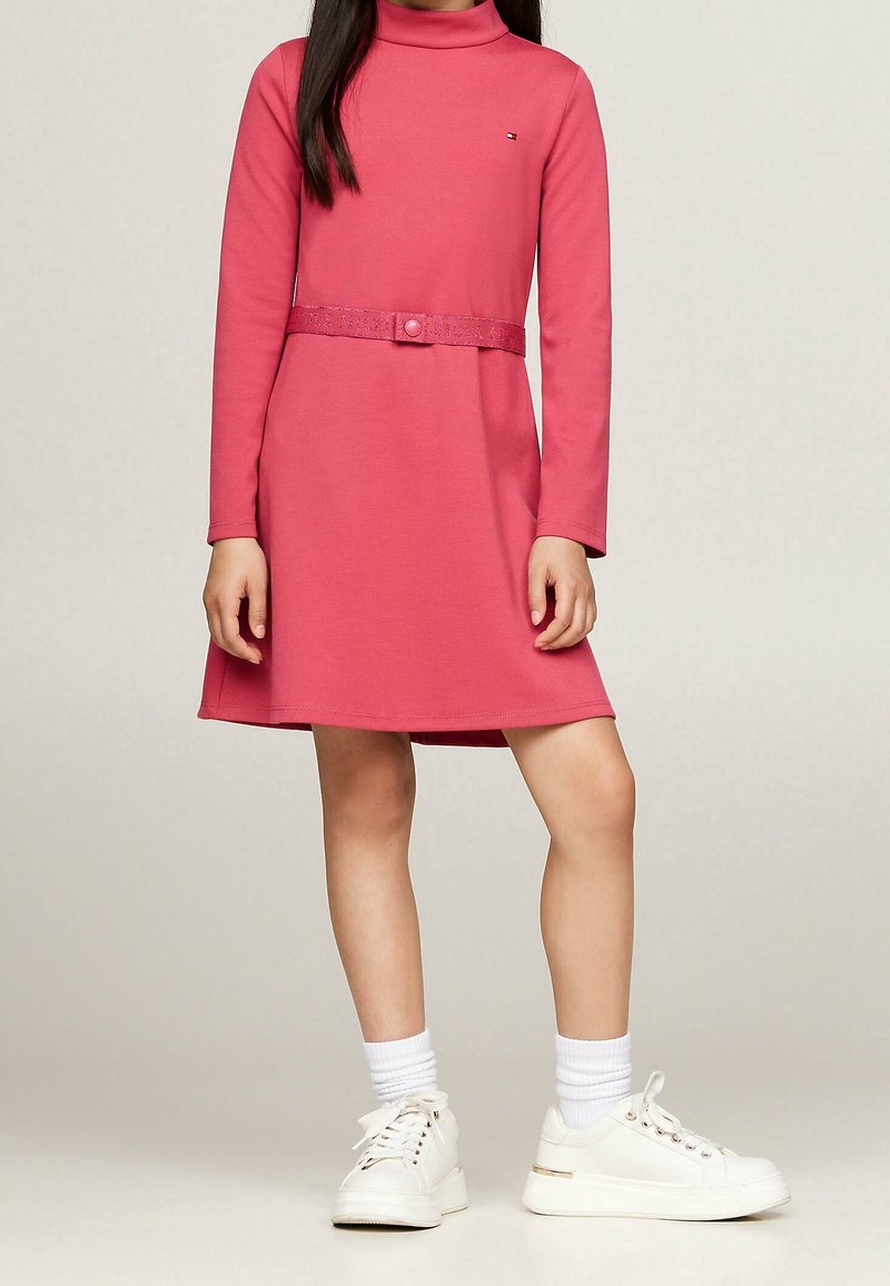 Różowa sukienka z długim rękawem z wysokim kołnierzem, z paskiem w talii i teksturowanym paskiem. Połączona z białymi sneakersami i prążkowanymi skarpetkami.