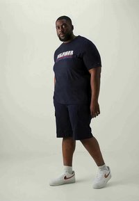 Man wearing navy graphic t-shirt, navy cargo shorts, white socks, and white Nike sneakers standing against plain background.