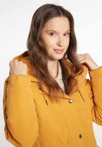 Žlutá zateplená bunda s kapucí, vybavená stahovacími šňůrkami, stříbrnými knoflíky a hladkým povrchem. Model nosí pod ní světlý top.
