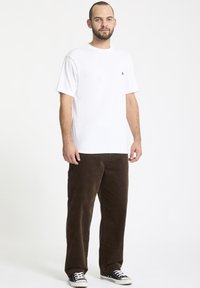 Hvid kortærmet t-shirt med et lille sort logo, kombineret med mørkebrune kordfløjl bukser og sorte sneakers, stående foran en ensfarvet baggrund.