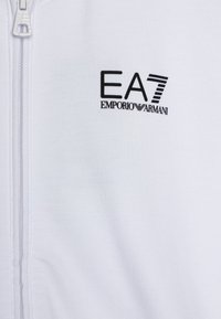 Balta ādas jaka ar struktūru un melnu izšūtu logo "EA7 EMPORIO ARMANI." Redzama rāvējslēdzēja detaļa.