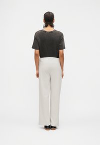 Άτομο με κοτσίδες cornrow στέκεται με την πλάτη προς τα εμπρός, φοράει μαύρο κοντό μπλουζάκι, άσπρο παντελόνι με φαρδιά πόδια και μαύρα παπούτσια σε λευκό φόντο.