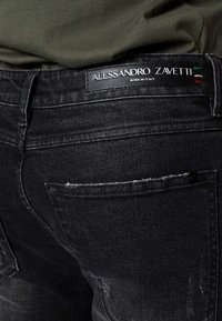 Vaqueros de mezclilla negros con textura desgastada, que presentan una etiqueta de cuero con la inscripción "Alessandro Zavetti" y una pequeña etiqueta en el bolsillo trasero.