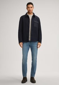 Veste en laine bleu marine avec col, fermeture à boutons sur le devant et poche sur la poitrine, portée par-dessus un pull gris clair et un jean bleu.