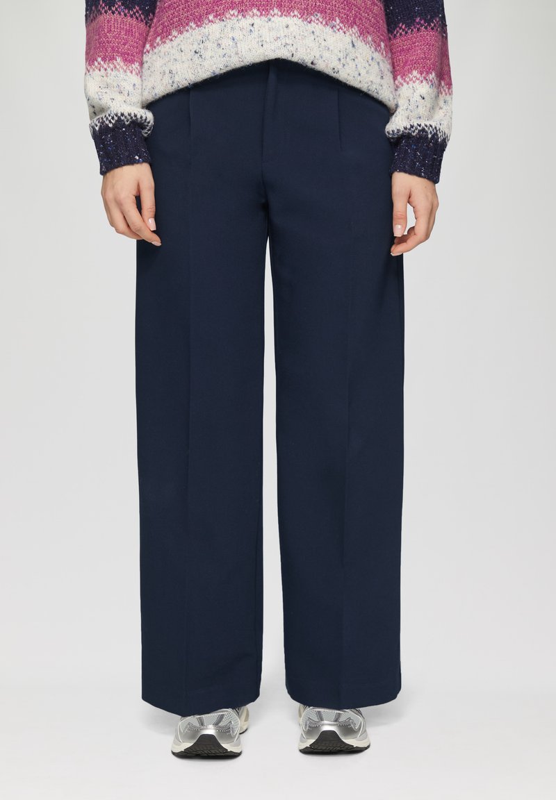 Marineblauwe wijde broek gemaakt van stof met een gladde textuur, met voorplooien en een rechte snit. Gecombineerd met zilveren sportieve schoenen.