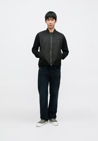 Crna bomber jakna, tamnoplave traperice i sivi teniski. Jakna ima zatvarač na patentni zatvarač i rebraste manžete, a model stoji na neutralnoj pozadini.