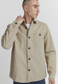 Beige langermet skjorte laget av bomull, med knappestolpe foran, brystlomme og svarte knapper som fremhever designet.