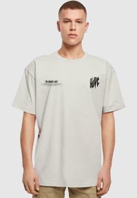 Oversized lysegrå T-shirt med korte ærmer, prydet med sort trykt tekst "HOPE" og "THE GUIDING LIGHT" på fronten. Blødt bomuldsstof.