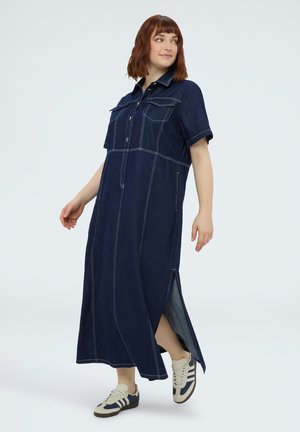 Kvinde iført en mørkeblå denim maxikjole med korte ærmer, forlommer, hvide syninger, sideslids og hvide sneakers med blå striber.