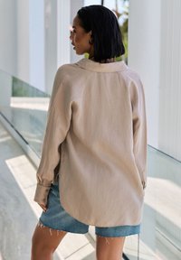 Beige long-sleeve shirt in lightweight fabric with a collar, curved hem, and buttoned cuffs. Paired with light blue denim shorts.