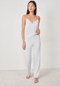 Vrouw in een lichtgrijze mouwloze top met kantafwerking en bijpassende losse broek, staande op blote voeten op een karpetten vloer tegen een effen muur.