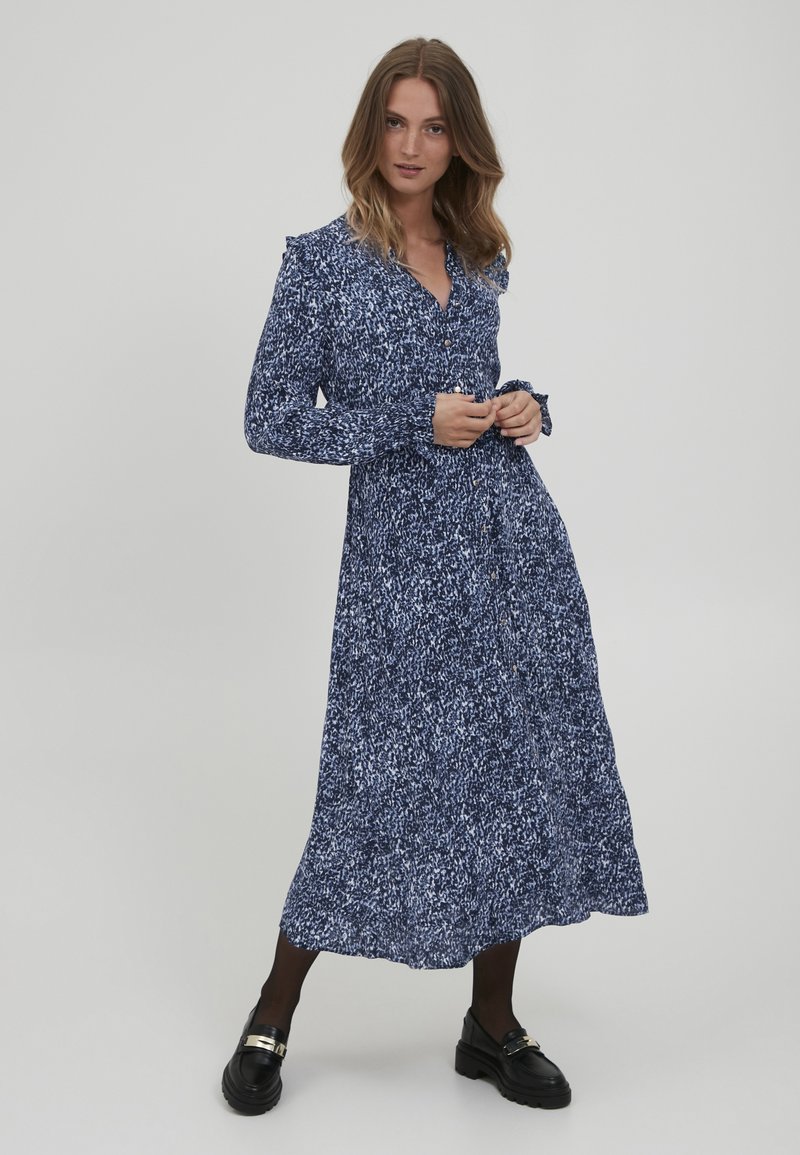 Robe longue bleu marine avec un motif texturé et moucheté, devant boutonné et accents de manches à volants, associée à des chaussures noires volumineuses.