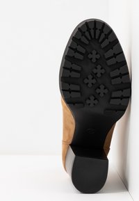 Semelle en caoutchouc noire avec des rainures profondes et un talon bloc sur une botte en daim beige, photographiée par le dessous sur fond blanc.