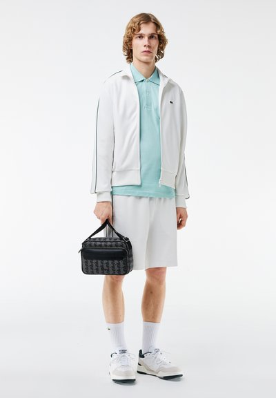 Veste zippée blanche avec des accents noirs, polo bleu clair, short blanc et baskets grises. Tient un sac noir à motifs avec une poignée.