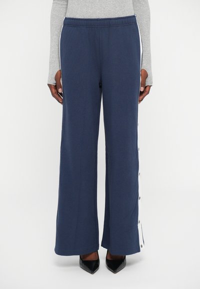 Pantaloni largi navy cu dungi albe cu capse pe laturi, purtați cu un top gri cu mâneci lungi și tocuri negre ascuțite.