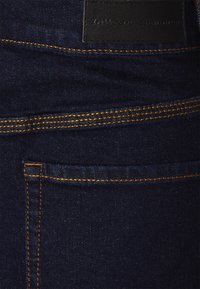 Jeans de mezclilla azul marino con costuras naranjas, que presentan un bolsillo trasero y una etiqueta negra. El material parece duradero con una textura suave.