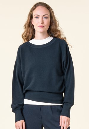 Frau mit langen lockigen Haaren, die einen locker sitzenden dunkelblauen Pullover über einem weißen Oberteil und passende dunkelblaue Hosen vor hellem Hintergrund trägt.