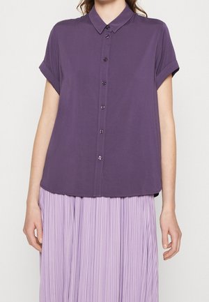 Femme portant une chemise violette à manches courtes, boutons fermés, avec manches retroussées et une jupe plissée violet clair sur un fond uni.