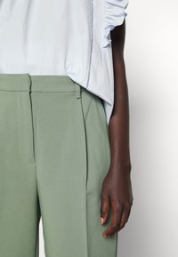 Chemise bleu clair à manches bouffantes, associée à un pantalon vert taille haute et ajusté. Une main visible repose sur la jambe du pantalon.