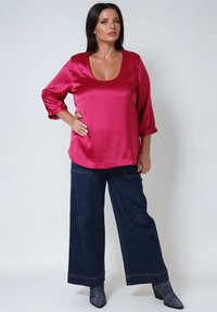 Woman with dark hair wears a magenta satin blouse with three-quarter sleeves, dark wide-leg jeans, and gray heeled boots, posing with hand on hip.