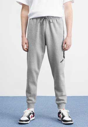 Person iført grå joggebukser med snøre og hvide, sorte og røde sneakers står på blåt tæppegulv.