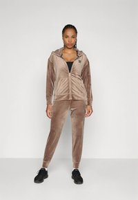 Καφέ βελούδινη φόρμα με φερμουάρ και τσέπες στο φούτερ με κουκούλ�α. Το παντελόνι στενεύει στον αστράγαλο. Συνδυάζεται με μαύρα αθλητικά παπούτσια.