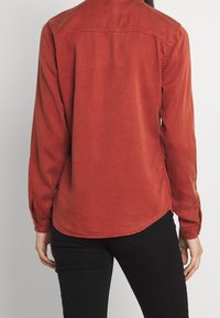 Camicia a maniche lunghe color ruggine con polsini abbottonati, orlo curvo e una consistenza morbida, vista dal retro.