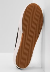 Kengän pohja, jossa on ruskea, tekstuurillinen kumituki ja valkoiset vahvistetut reunat valkoista kulmataustaa vasten.