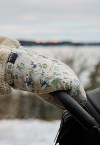 Hånd iført vante med blomsterprint og fleeceforing, der griber et sort klapvognshåndtag udendørs i koldt vejr.