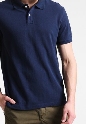 Mies, joka pukeutunut tummansiniseen, lyhythihaiseen poolopaitaan, jossa on kaksi nappia, ja oliivinvihreisiin housuihin, seisoo käsi taskussa.