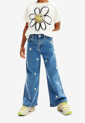 White t-shirt with a large daisy graphic and smiley face; blue jeans with embroidered white daisies; beige sneakers with bright green accents.