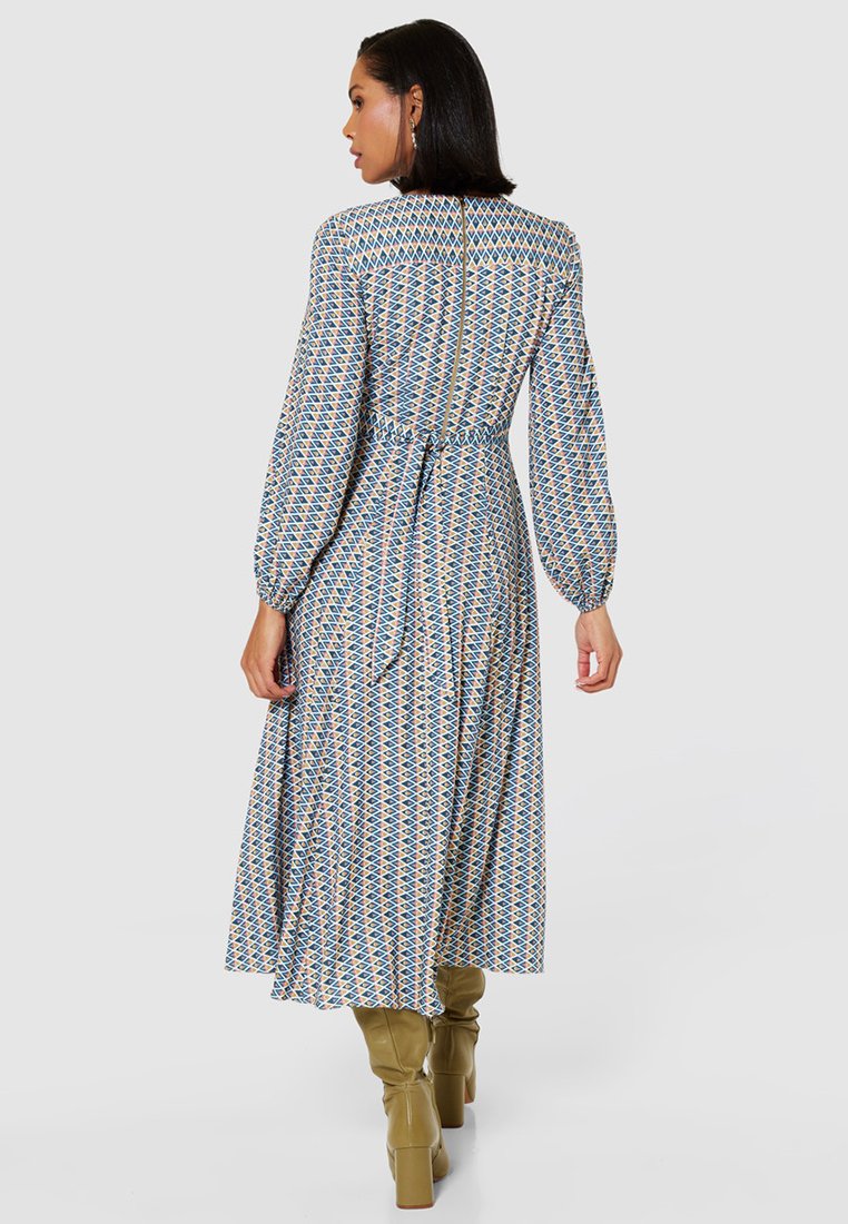 Sukienka z długim rękawem, we wzory, z niebieskim, żółtym i czerwonym geometrycznym wzorem, dopasowana w talii, z wiązanym paskiem. Połączona z zielonymi kozakami do kolan.