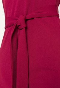 Gepunktetes Kleid aus tiefmagenta Stoff mit glatter Textur und einer gebundenen Knotendetails. Das Design betont eine schmeichelhafte Silhouette.