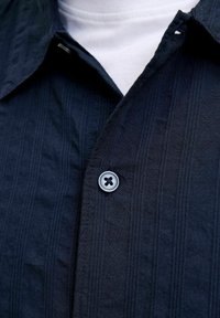 Gros plan d’une chemise bleu marine foncé boutonnée avec des rayures verticales discrètes portée sur un sous-vêtement blanc.
