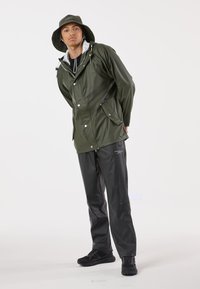 Groene waterdichte jas met capuchon, zilverkleurige drukknopen en zakken, gecombineerd met zwarte waterdichte broek en zwarte sportschoenen.