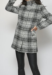 Černobílý kostkovaný kabát z texturované látky s vysokým límcem, dlouhými rukávy a bočními kapsami; nošený přes černé přiléhavé kalhoty.