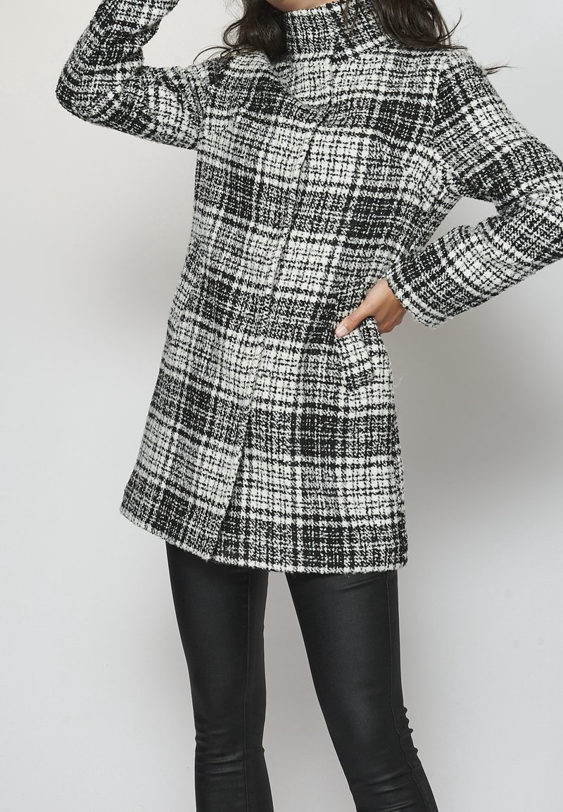 Černobílý kostkovaný kabát z texturované látky s vysokým límcem, dlouhými rukávy a bočními kapsami; nošený přes černé přiléhavé kalhoty.