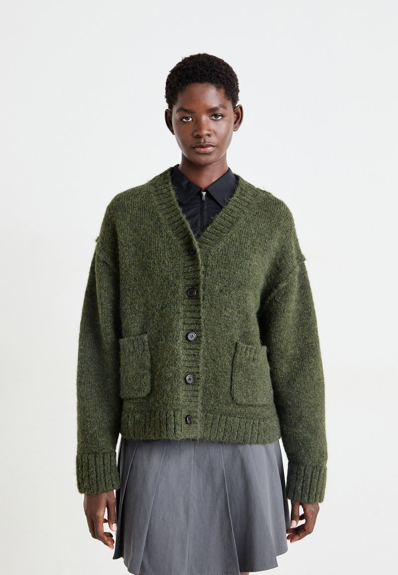 Groene wollen cardigan met een V-hals, grote knopen, twee voorzakken en geribde manchetten, gedragen over een zwart shirt en een grijze plissé rok.