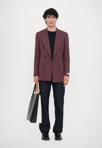 Burgunder Doppelreiher-Blazer, dunkles T-Shirt, dunkle Jeans, schwarze Schuhe, hält eine schwarz-weiße Tragetasche. Glattes Material, taillierte Passform.