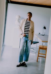 Bežinė švarkas ant juodai baltų dryžuotų marškinių, plačios kojos mėlyni džinsai, juodi batai, minimalistinis interjeras ir užnugaryje biusto statula.