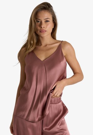 Donna con top in raso mauve senza maniche e pantaloncini abbinati, con una mano in tasca, su uno sfondo chiaro e uniforme.